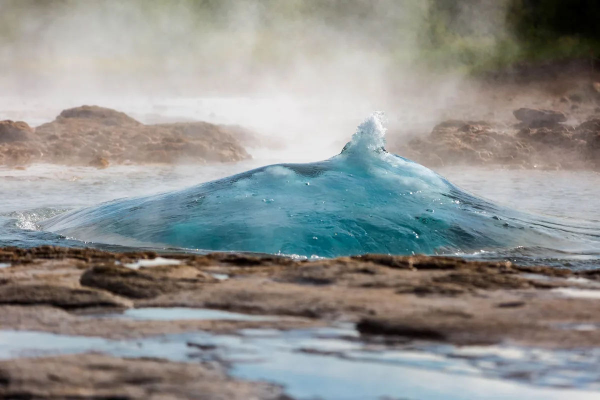 Close-up of a vibrant blue geyser bubbling up with steam in a rocky landscape.
