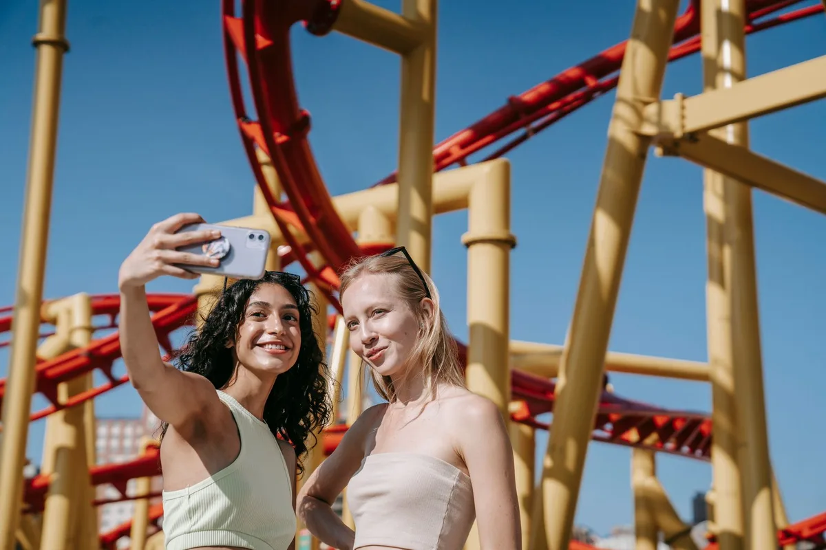 Two young women taking a selfie in front of a roller coaster at an amusement park.