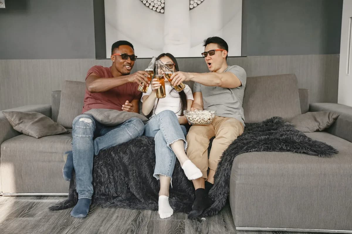 Three friends relax on a sofa, clinking beer bottles and sharing popcorn during a movie night indoors.