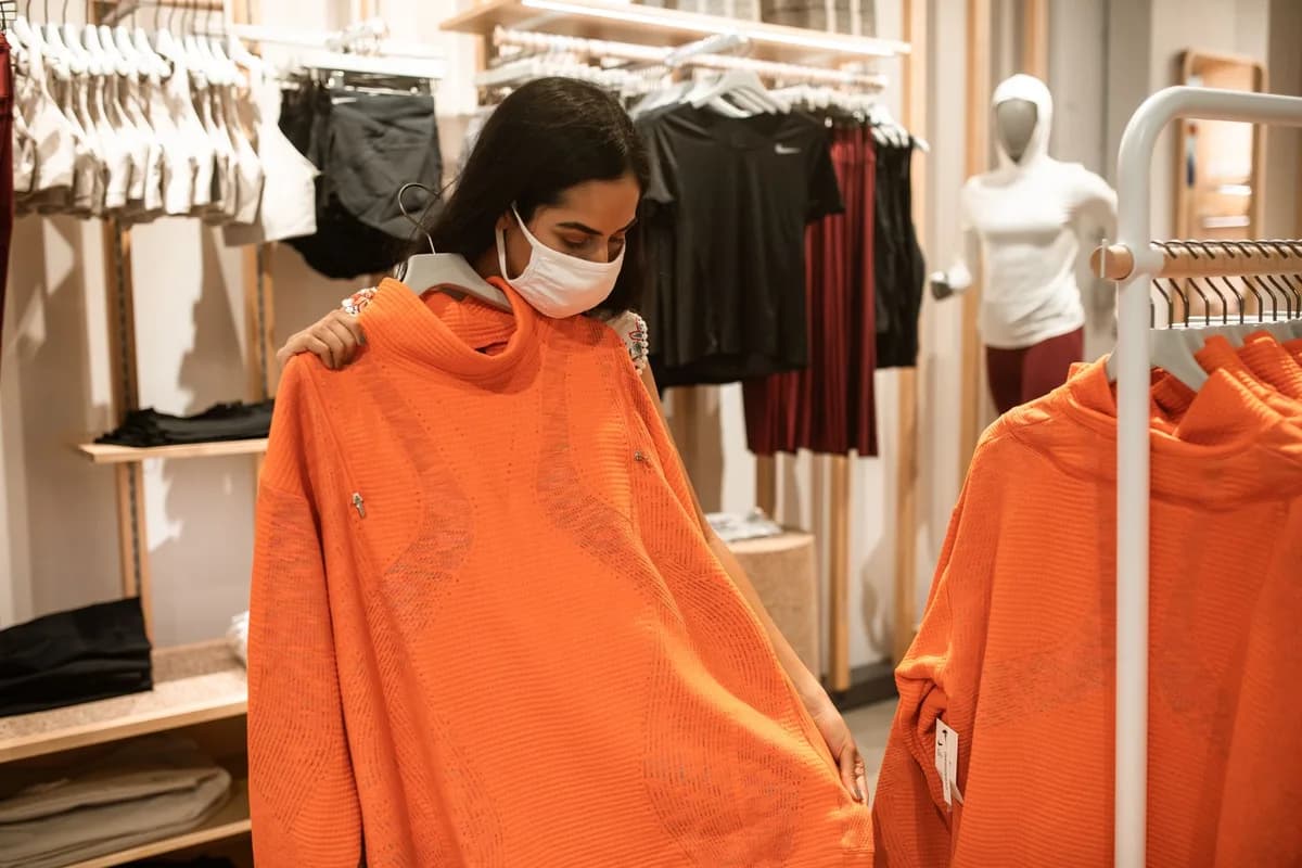 Woman wearing a face mask examines an orange sweater in a clothing store.