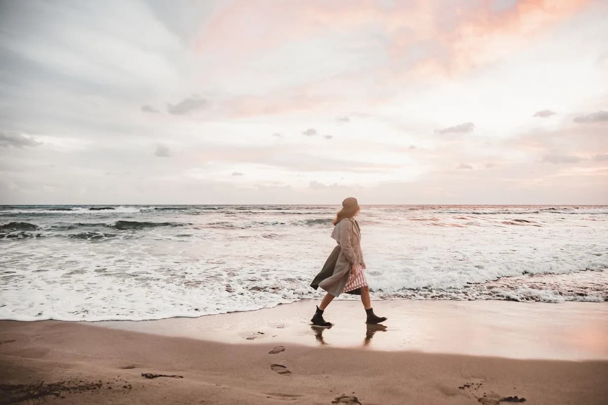 A woman walks along a sandy beach with a stunning sunset and gentle waves creating a serene atmosphere.