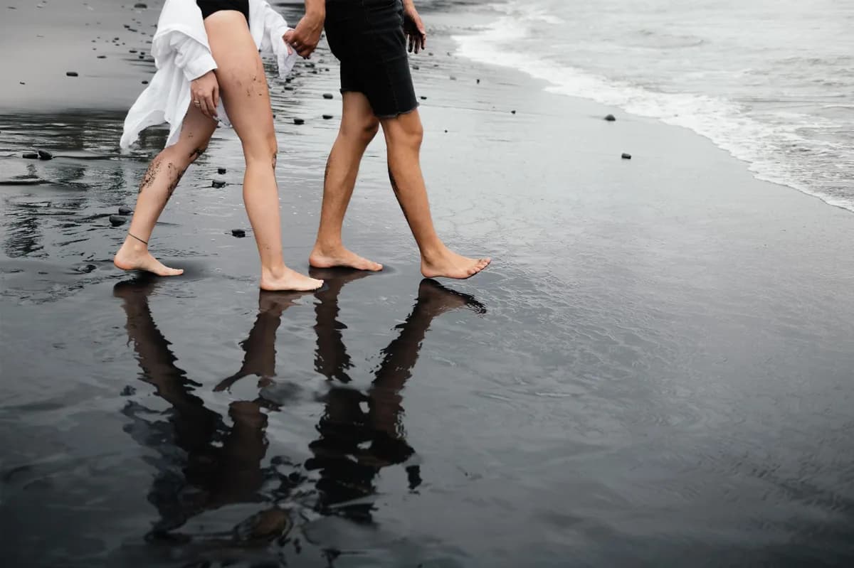 A barefoot couple walking along a black sand beach, reflecting off wet sand.