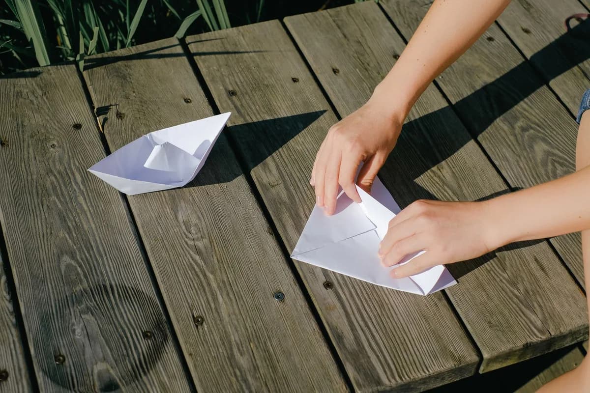 Close-up of hands folding paper boats on a wooden deck, perfect for creative and leisure-themed concepts.