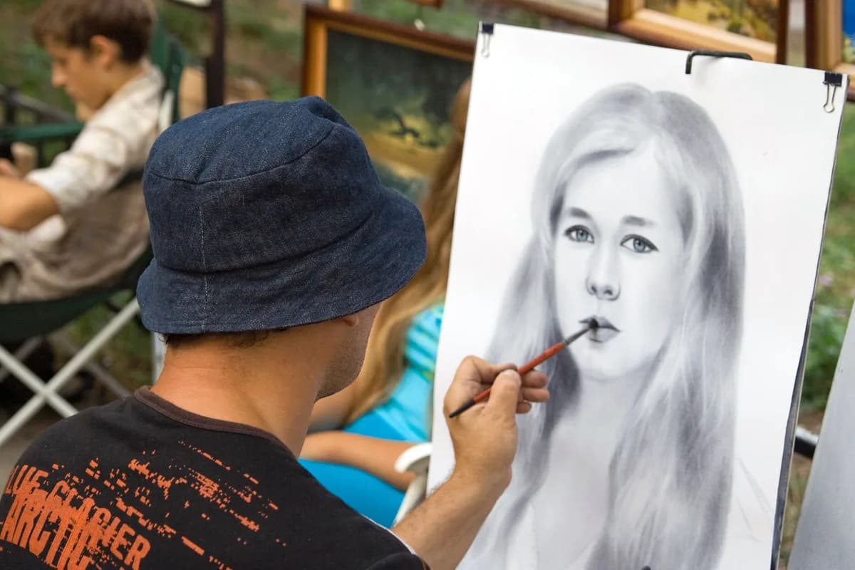 An artist drawing a realistic portrait outdoors surrounded by paintings.