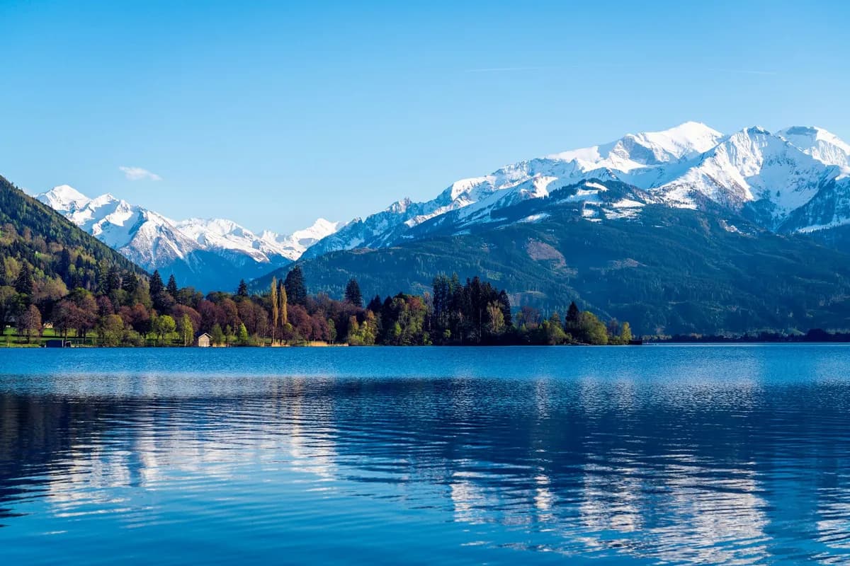 Snow-covered peaks mirrored in a tranquil lake under a clear blue sky, offering a breathtaking view of nature's beauty.