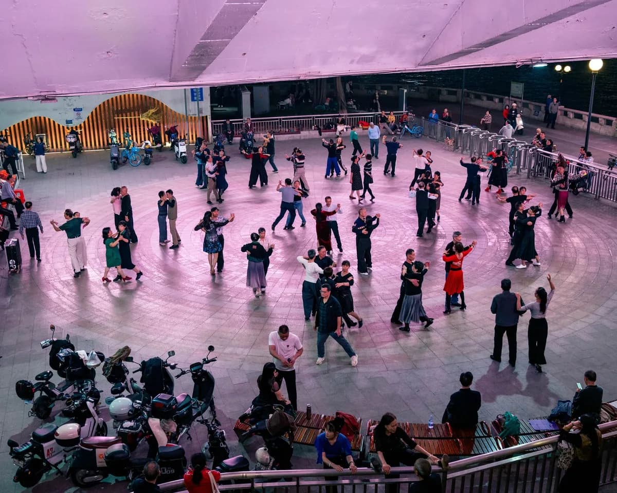 A large group of people dancing in an outdoor urban plaza under purple lights during the evening.
