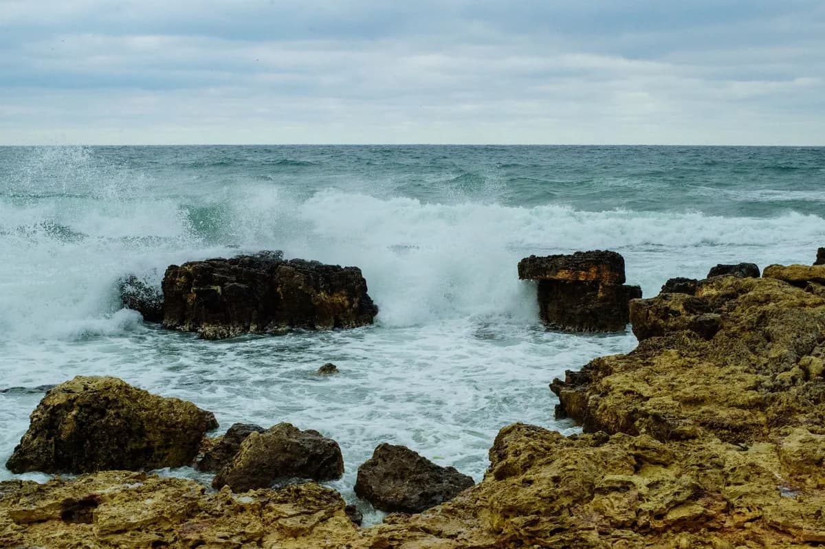 Dramatic view of ocean waves crashing on rocks at the shoreline under a cloudy sky.