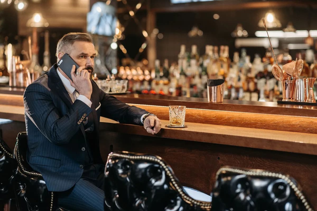 Elegant businessman in a suit making a phone call at a luxury bar with a glass of whiskey.