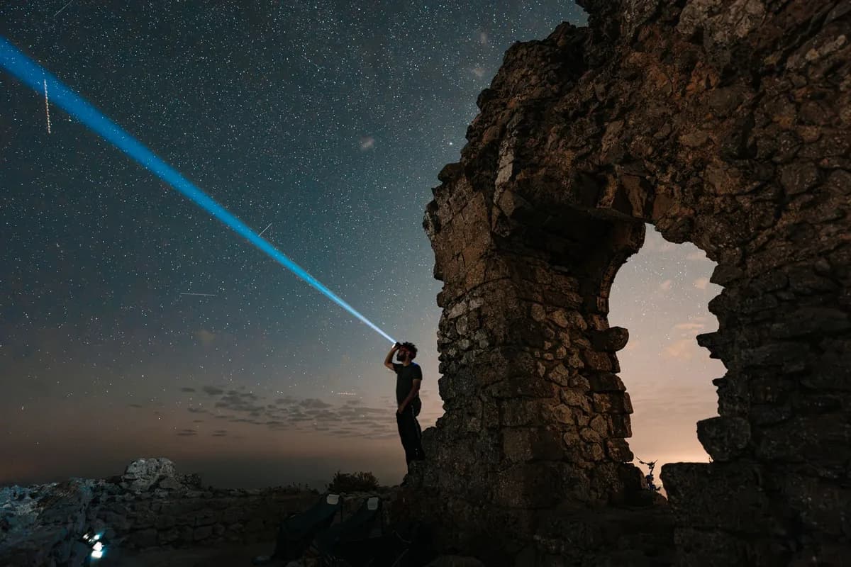 A person shines a flashlight at the star-filled sky by ancient ruins at night.