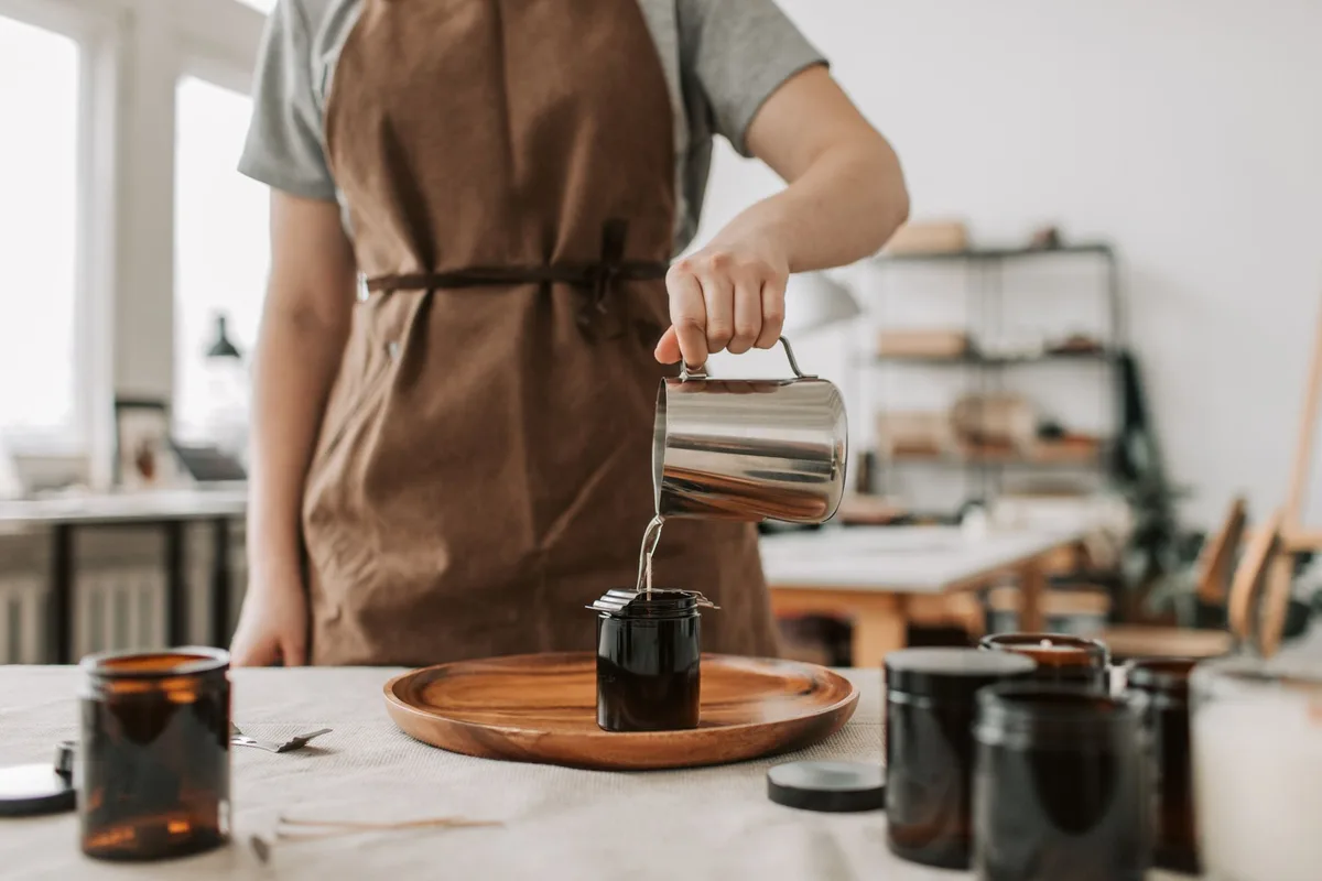 Person pouring wax into glass jars, creating handmade candles in a cozy workshop setting.