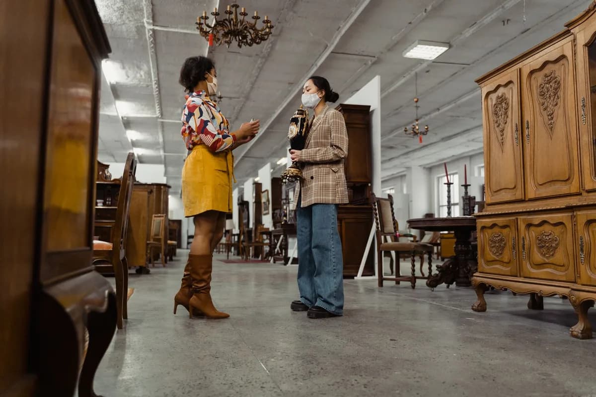 Two women wearing masks browsing antique furniture in a vintage store setting.