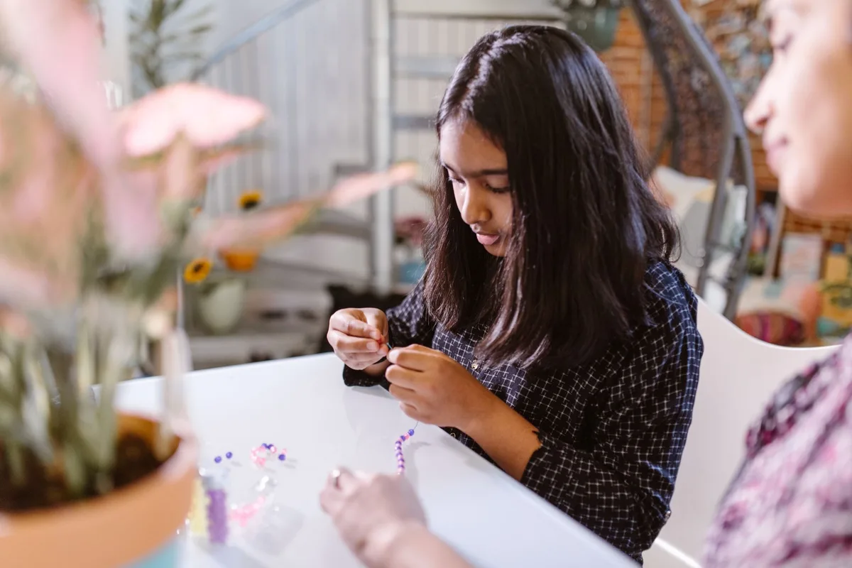 A young girl concentrating on crafting a beaded bracelet at a table indoors.