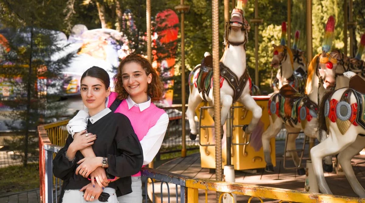 Two young women smiling and embracing near a carousel in an amusement park.