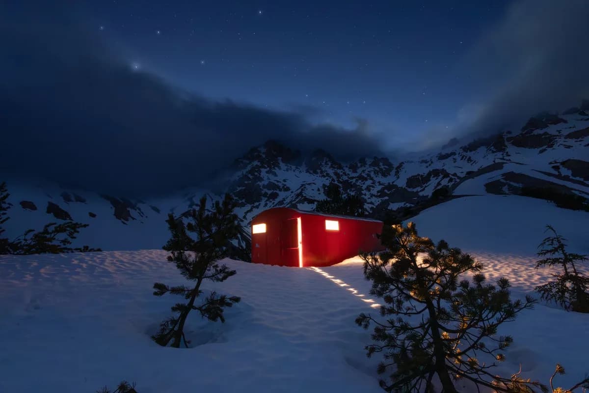 Red mountain hut illuminated under a starry night sky, surrounded by snow and trees.