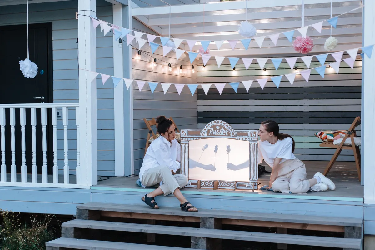 Two women enjoy an outdoor shadow puppet show on a porch decked with decorative pennants.