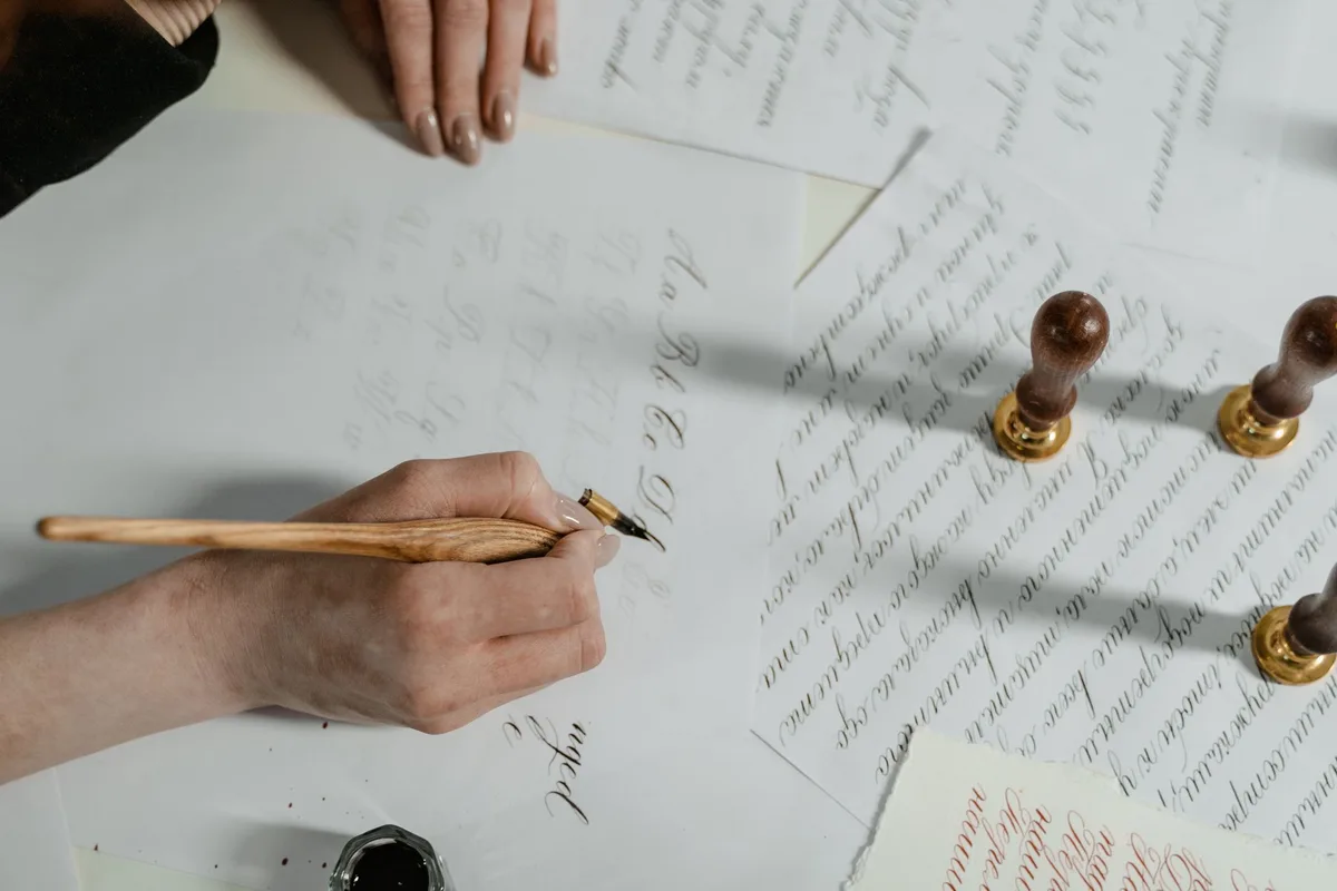 Close-up of a hand practicing calligraphy with a wooden pen, surrounded by ornate paper and wax seals.