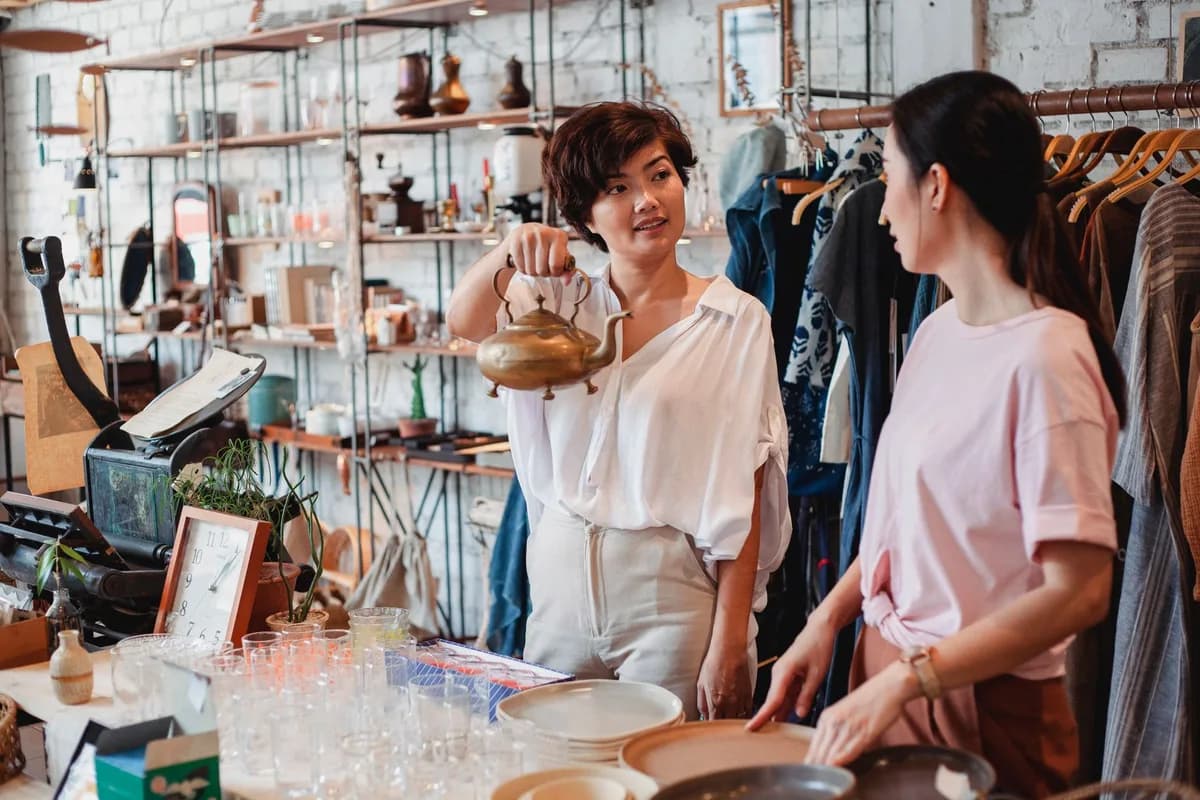 Young ethnic female shopper demonstrating metal kettle to partner while interacting near table with dishware in store and looking at each other