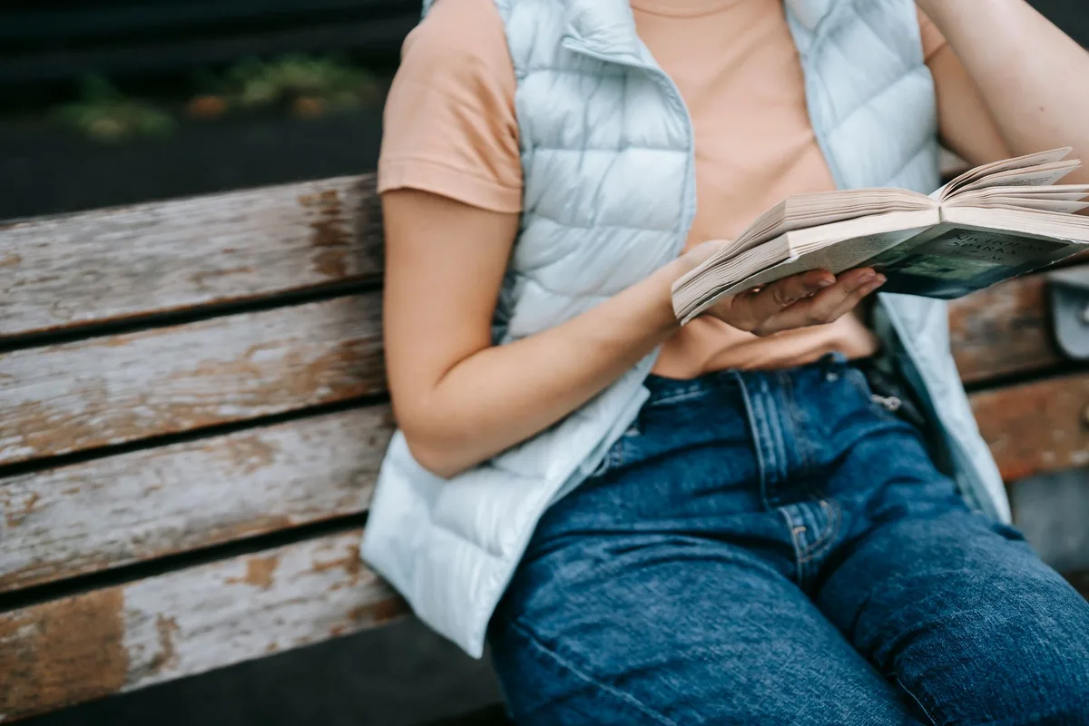 Crop faceless female in casual clothes sitting on aged wooden bench on street and reading book in daytime