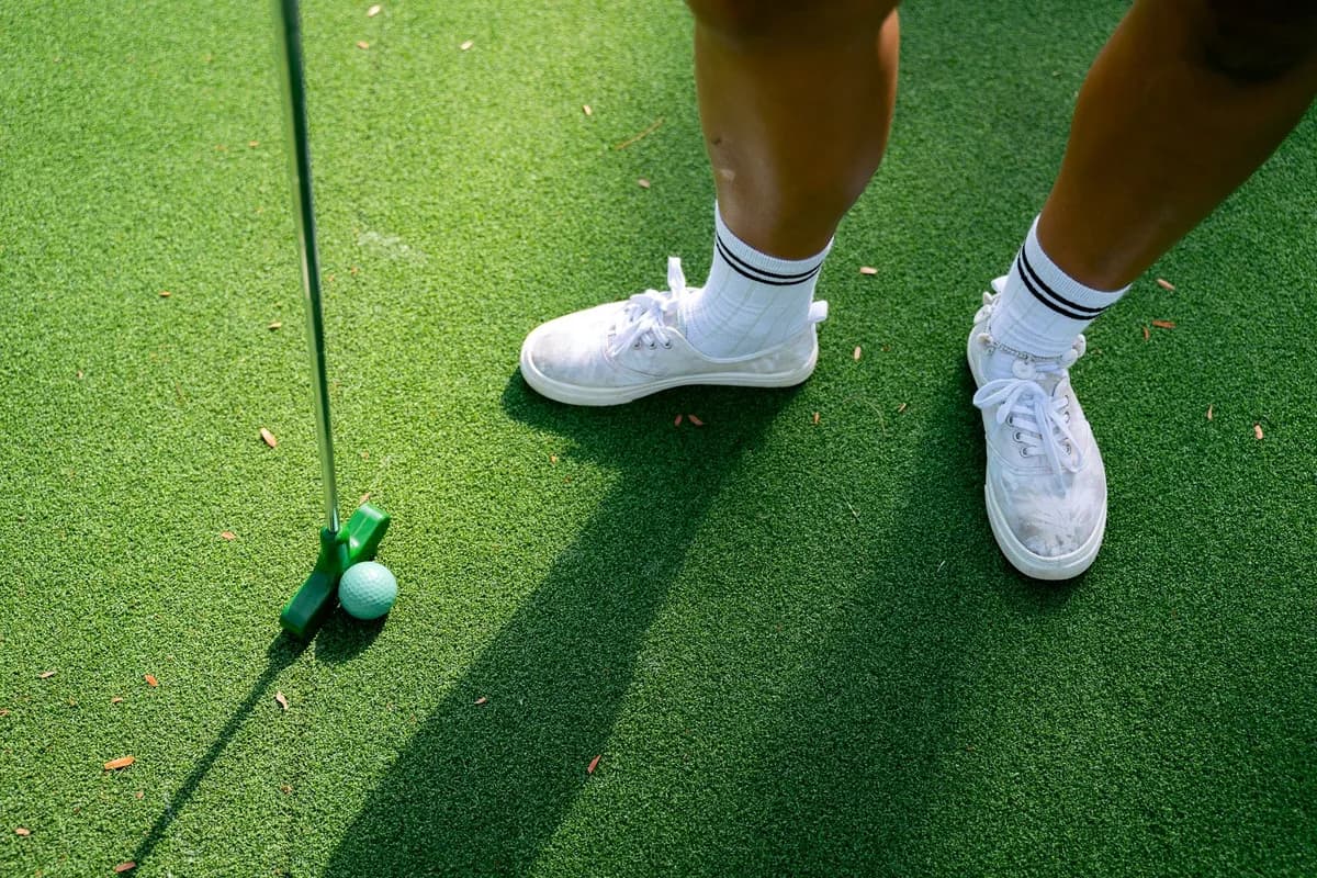 A person in white sneakers standing with a putter and golf ball on a green putting surface.
