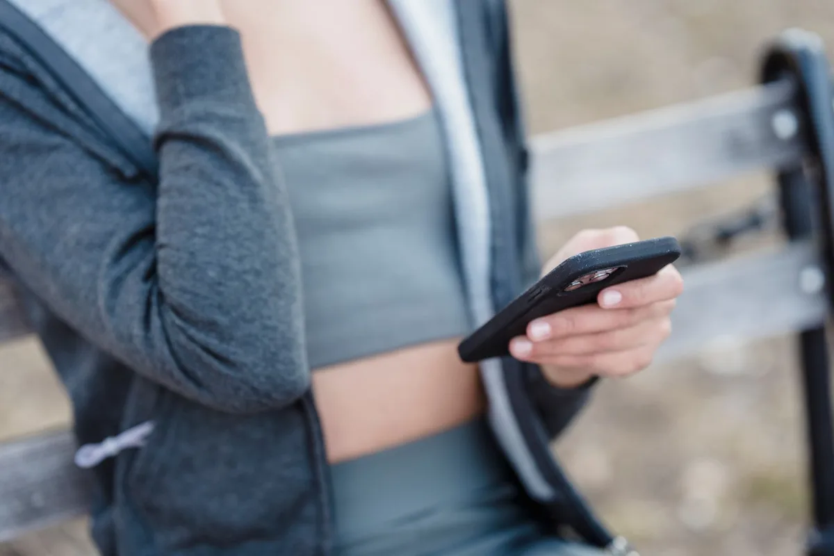 Crop unrecognizable slim female in sportswear text messaging on cellphone while sitting on bench on street against blurred background during