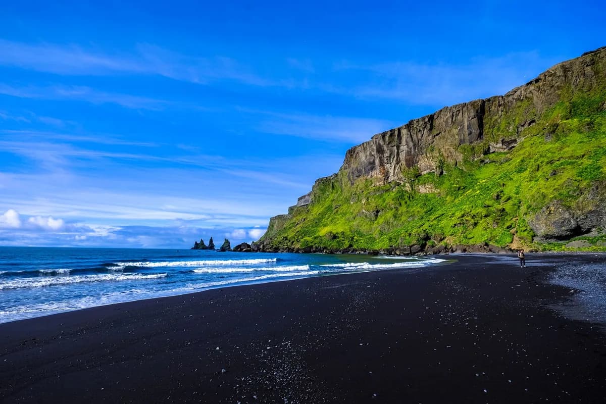 A black-sand beach beneath green cliffs and a blue sky.