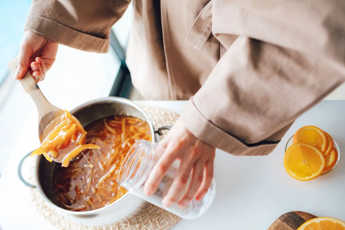 Hands adding marmalade into a jar from pot, with sliced oranges on the side.