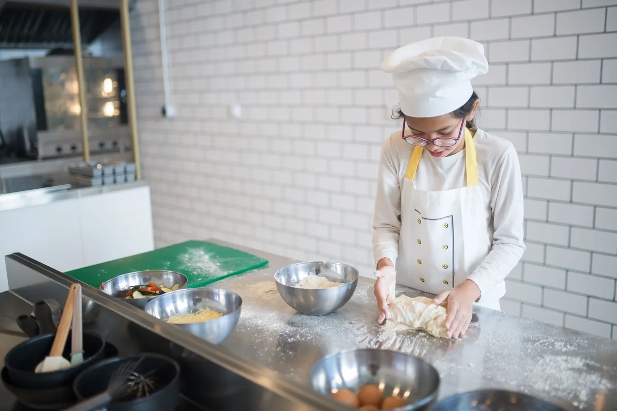 A young girl in a chef's hat kneads dough in a contemporary kitchen setting.
