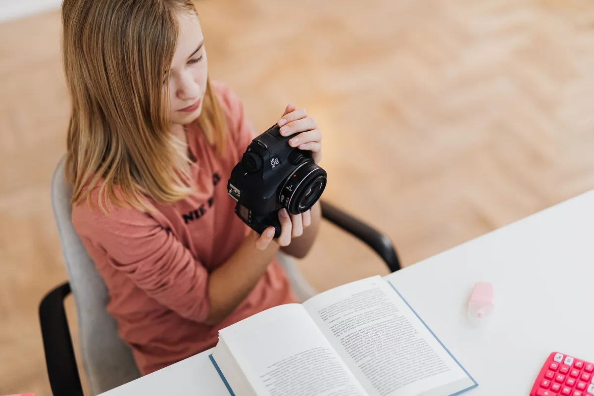 A young woman handling a DSLR camera while reading a book at a modern indoor setting.
