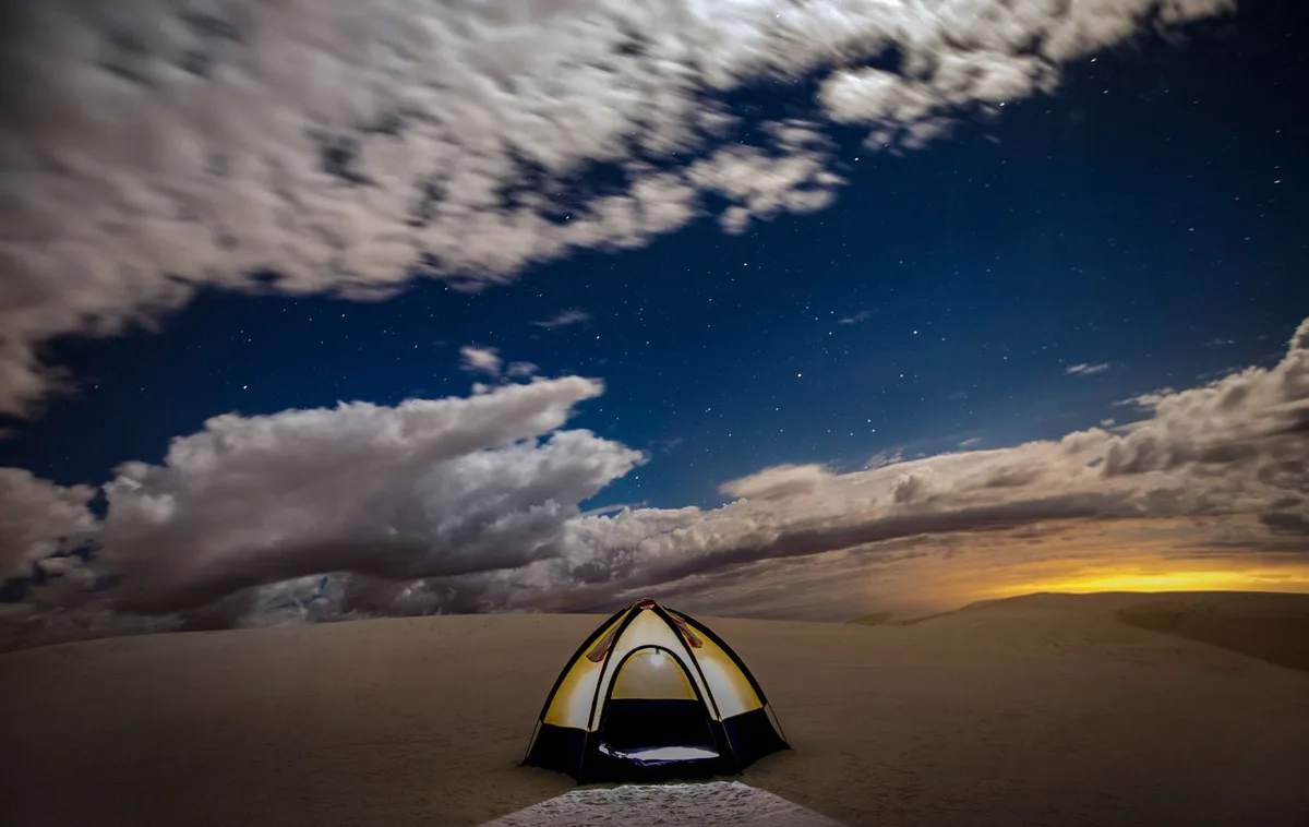 A solitary tent under a starry night in a vast desert, with dramatic skies and distant sunset glow.
