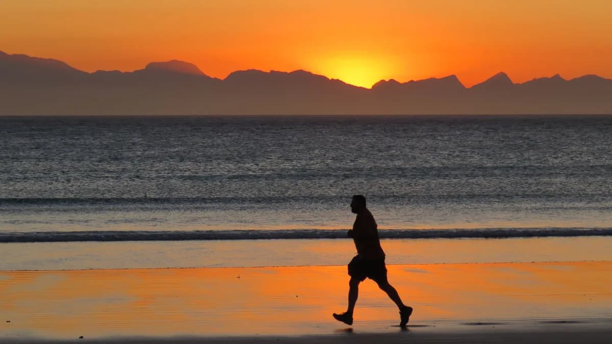 Silhouette of a man jogging along the shore at sunset with mountains in the distance.