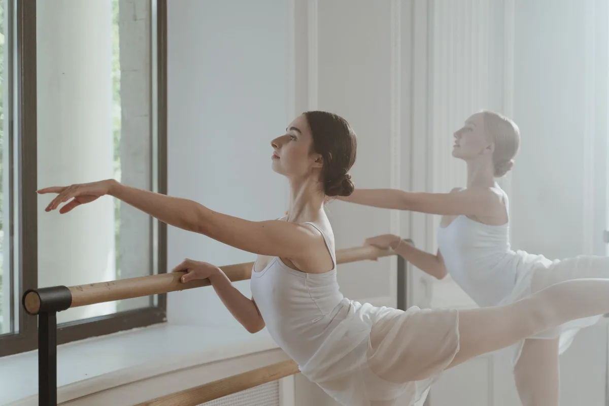 Elegant ballerinas in white leotards practicing ballet at a barre in a sunlit studio.