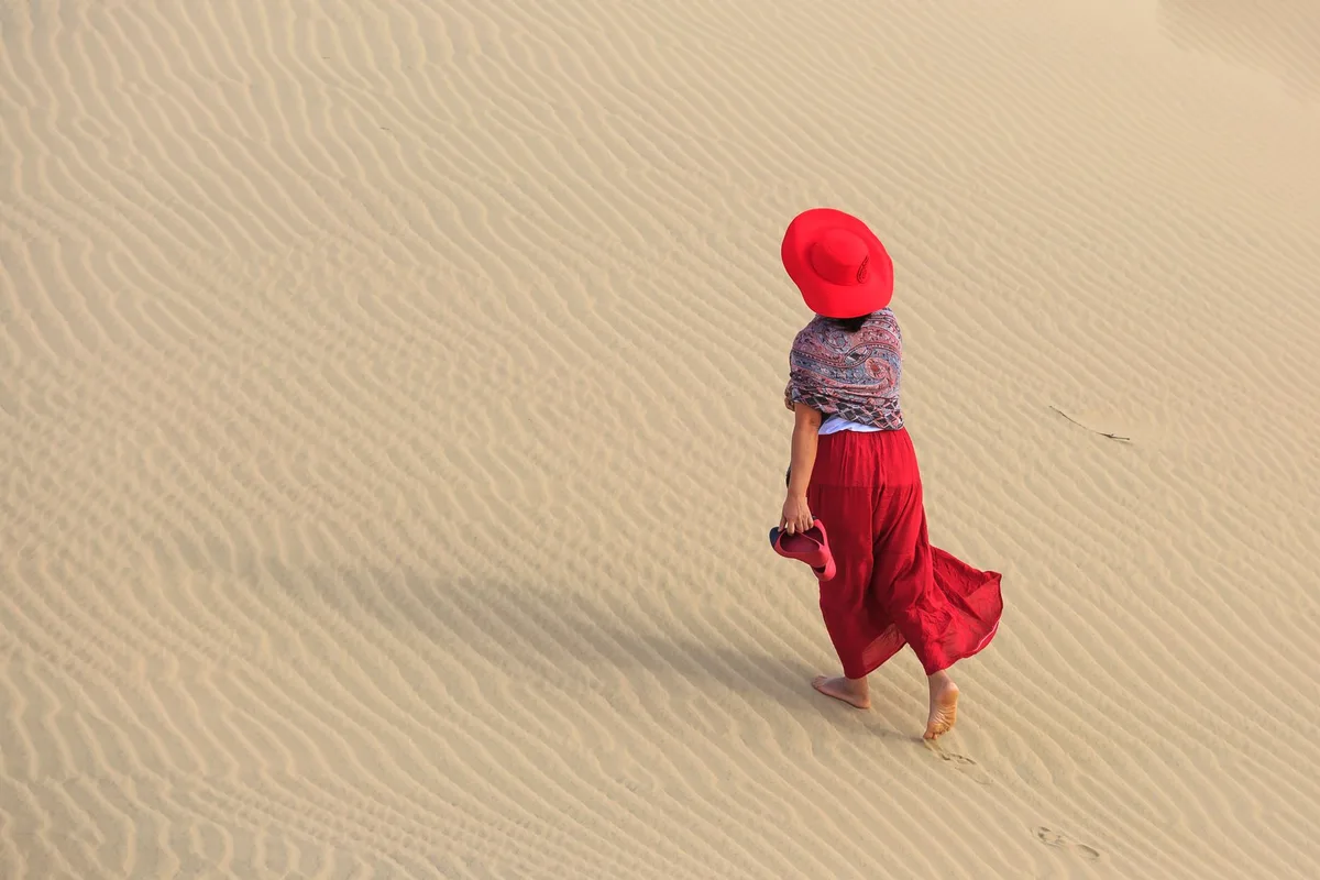 A woman in vibrant red attire walks barefoot across expansive sand dunes, creating a scenic desert landscape.