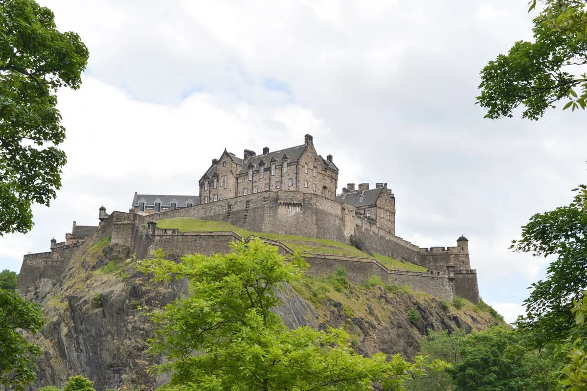 View of the iconic Edinburgh Castle perched on Castle Rock, framed by trees under a cloudy sky.