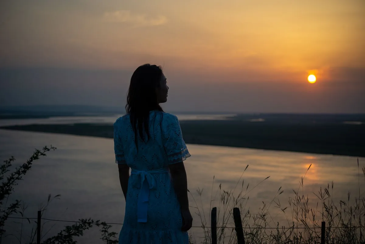 A serene silhouette of a woman in a dress overlooking a sunset by a river.