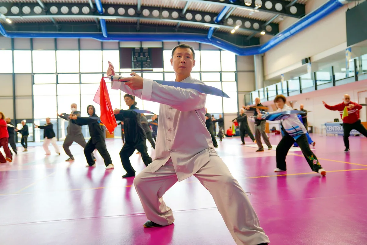 Group practicing Tai Chi with swords indoors, led by an instructor on a pink floor.