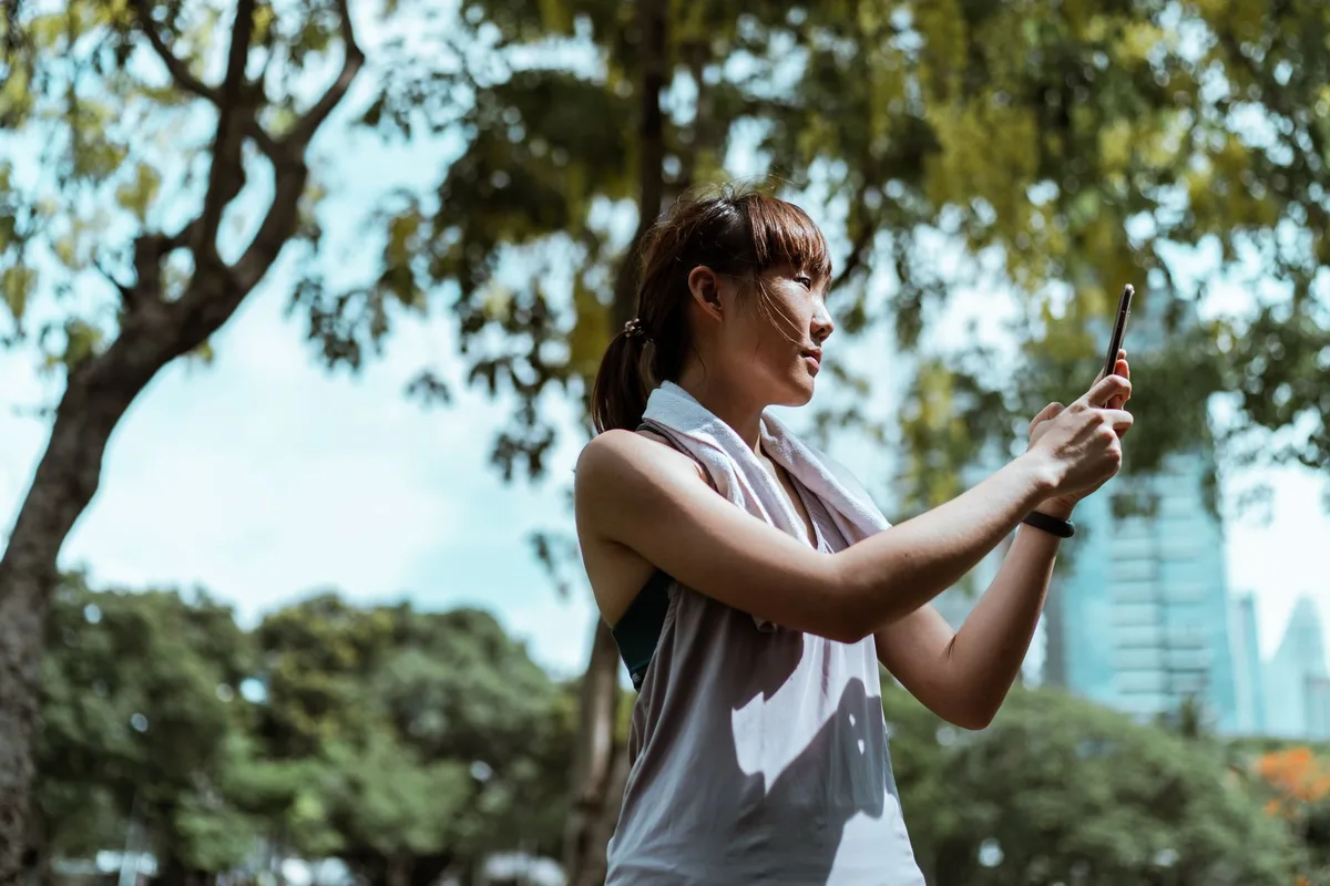 From below side view of slender Asian female athlete in sports clothes with towel taking selfie on mobile phone while standing near trees under sky and looking away
