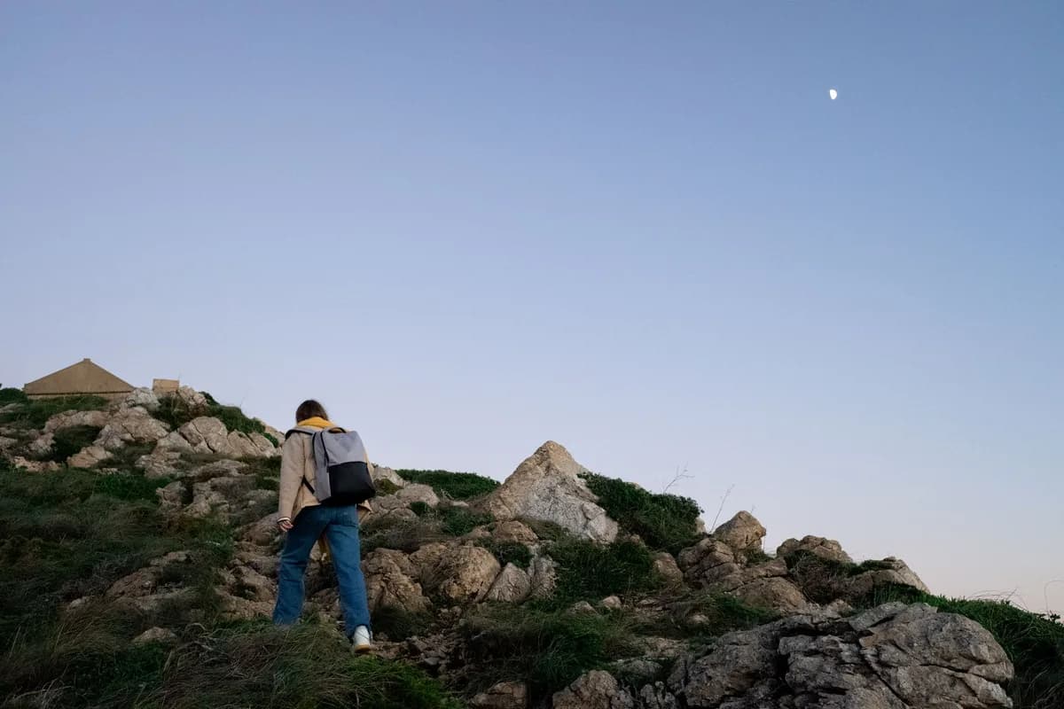 A woman hikes uphill on a rocky mountainside under a clear evening sky with the crescent moon visible.