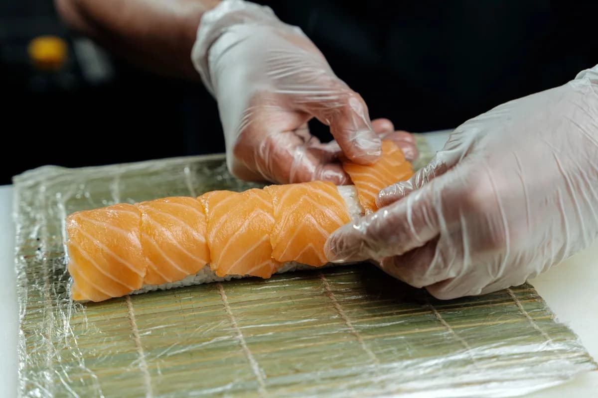 Close-up of chef's hands in gloves crafting salmon sushi roll, showcasing sushi preparation.