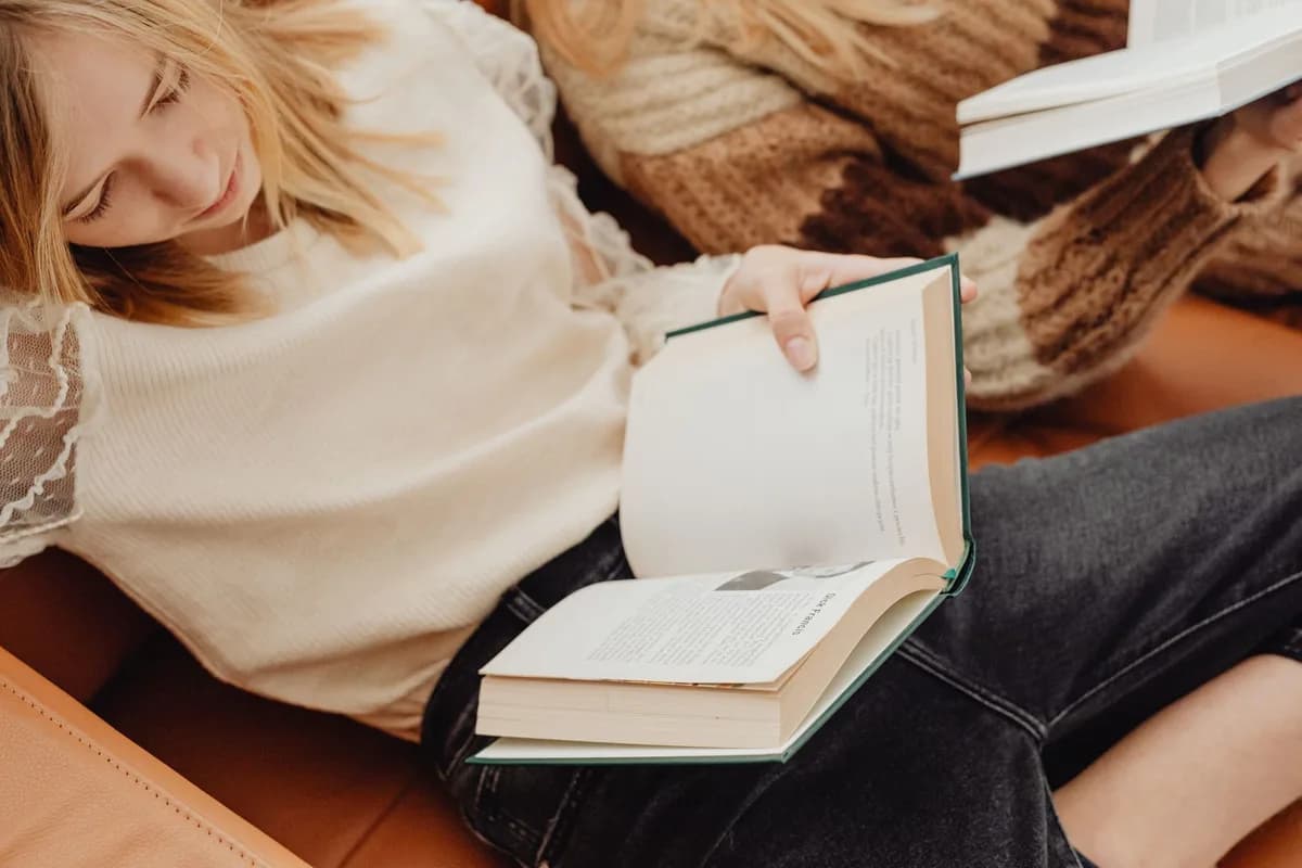 Two young women relaxing on a couch while reading books indoors.