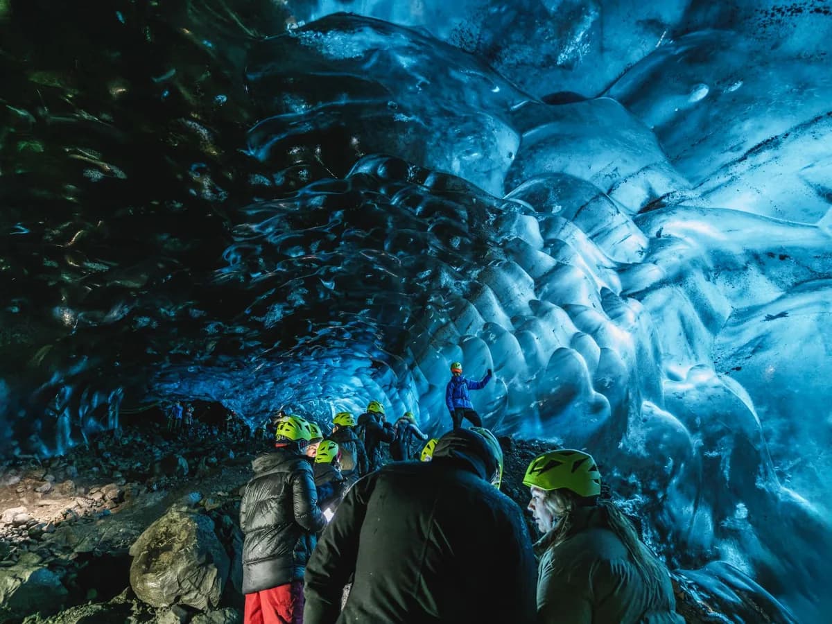 Group of explorers in an illuminated ice cave with vibrant blue hues, wearing helmets and winter gear.