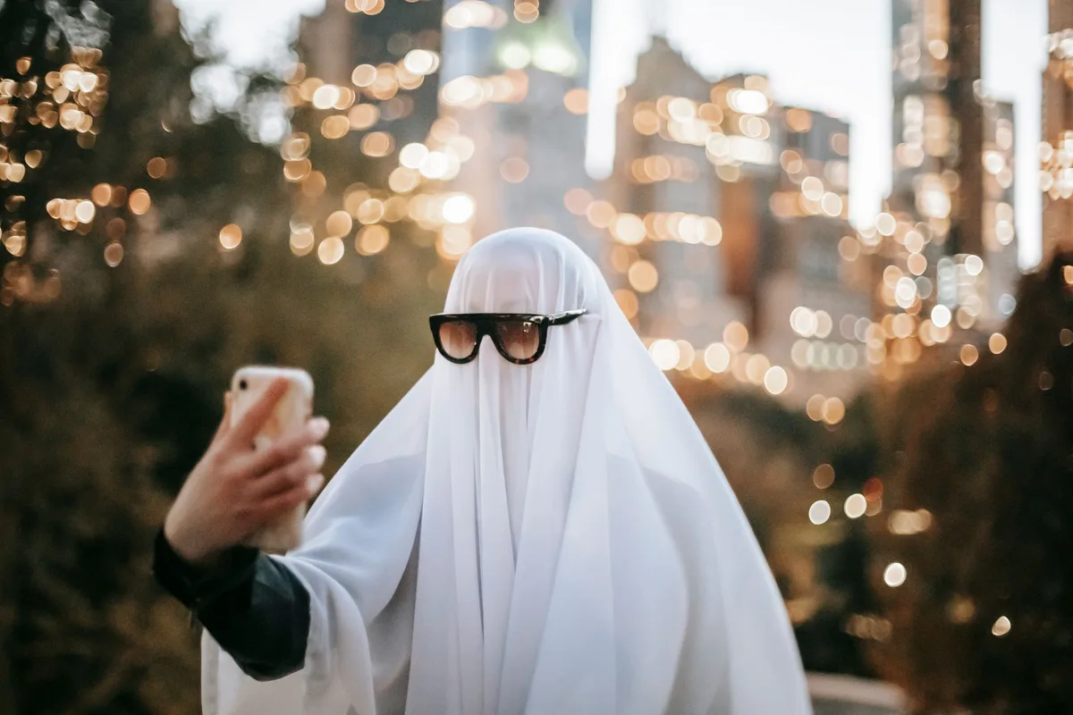 Anonymous person wearing ghost costume and sunglasses taking self portrait on smartphone while standing on street with trees and skyscrapers on blurred background during Halloween