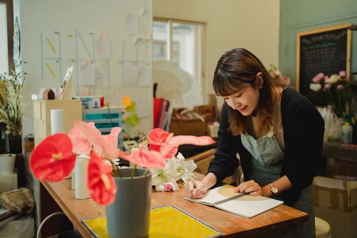 Calm florist in apron standing at table with flowers and writing in notepad in flower shop