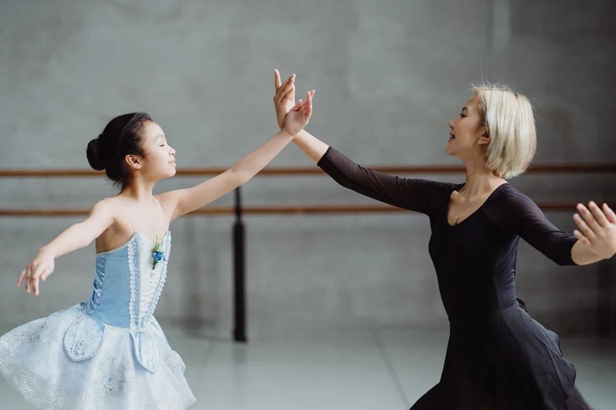 Side view of graceful Asian girl wearing tutu standing near teacher in black outfit with raised arms in ballet studio