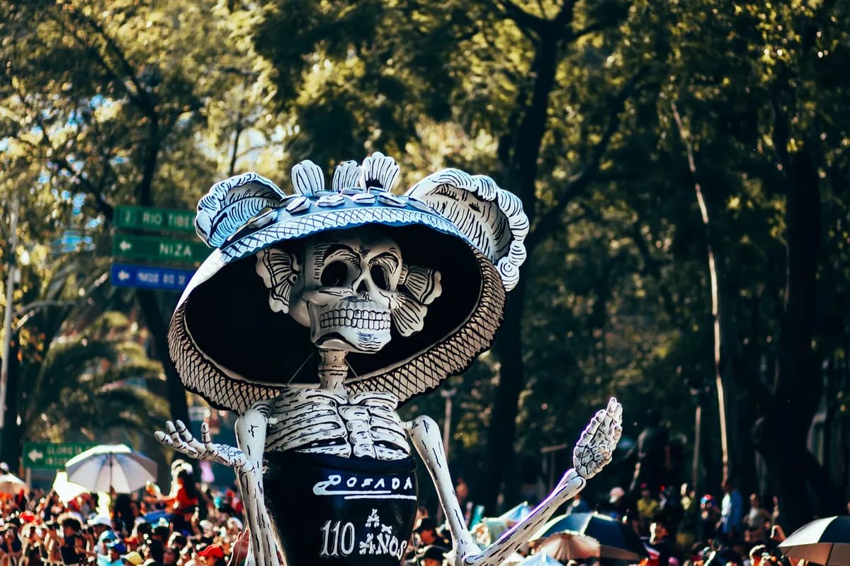 A colorful Day of the Dead parade with a giant skeleton sculpture in a lively city street.