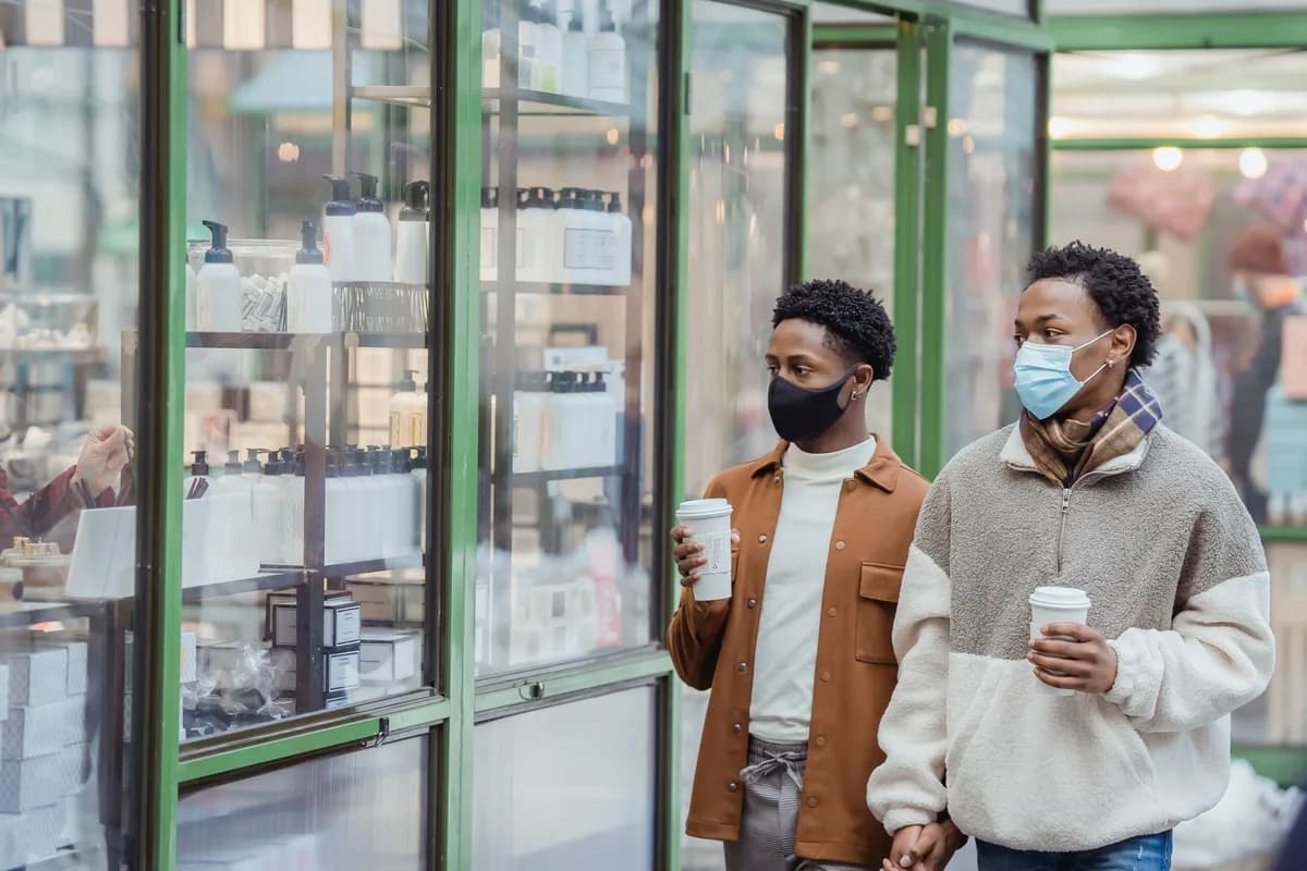 African American homosexual couple with takeaway beverages in medical masks holding hands while strolling on street near glass case of store in city