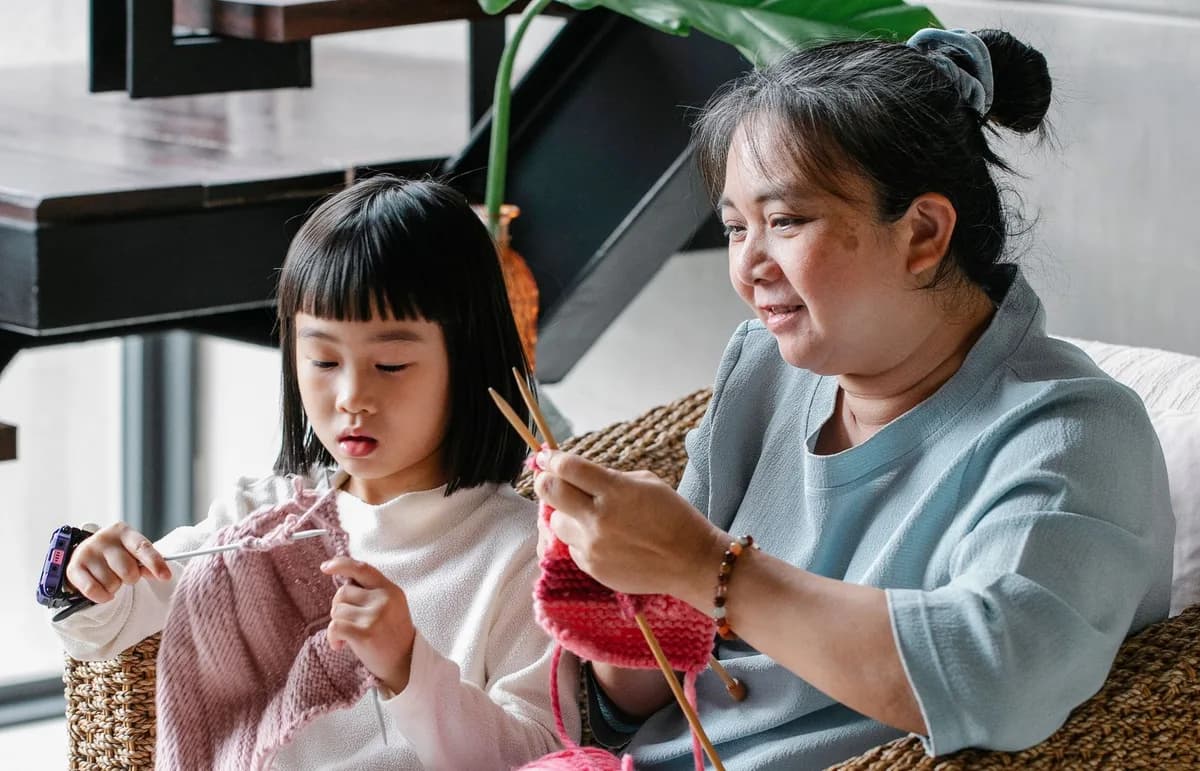 Happy mature Asian woman with little granddaughter sitting in cozy armchair and knitting with needles