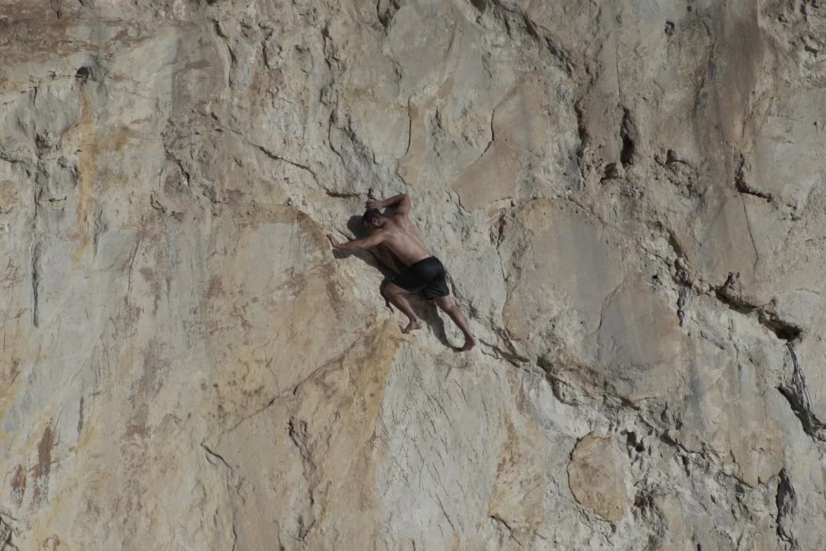 Adult male rock climber ascending a steep, rugged cliff outdoors in daylight.