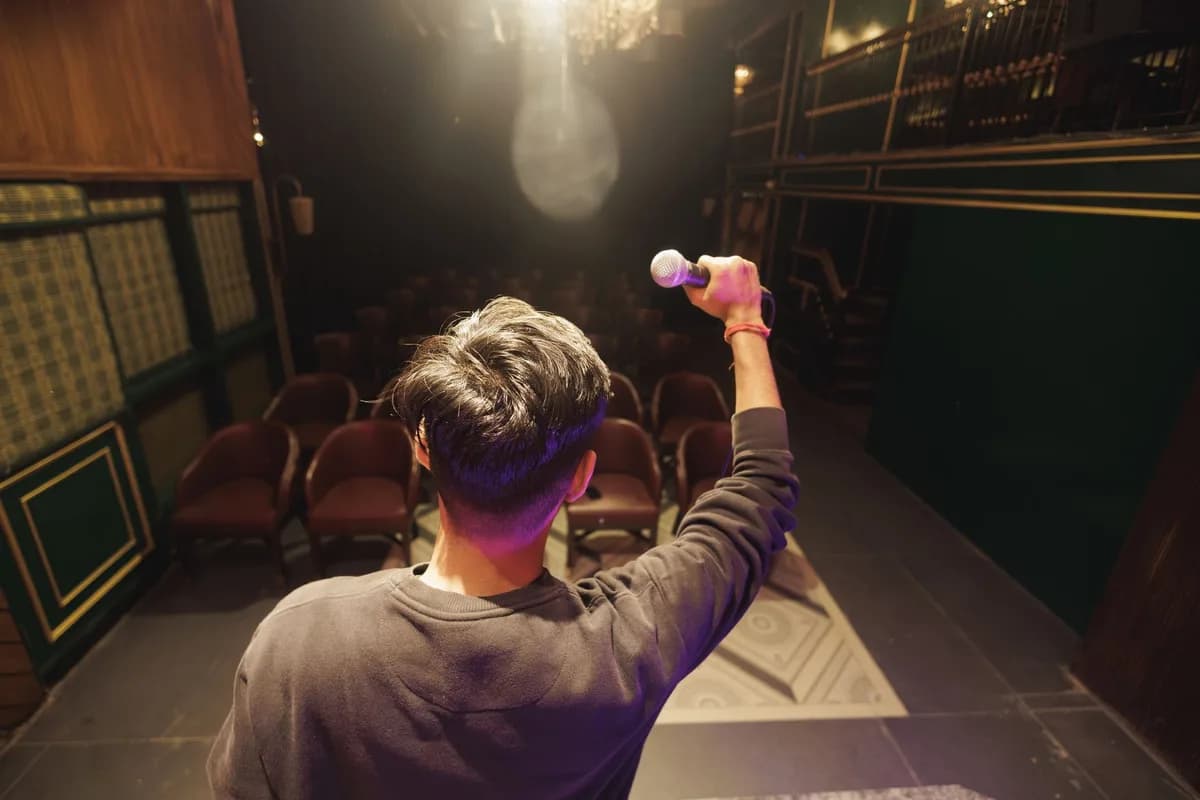 A young man on stage facing seats, holding a microphone, preparing for a performance in an auditorium.