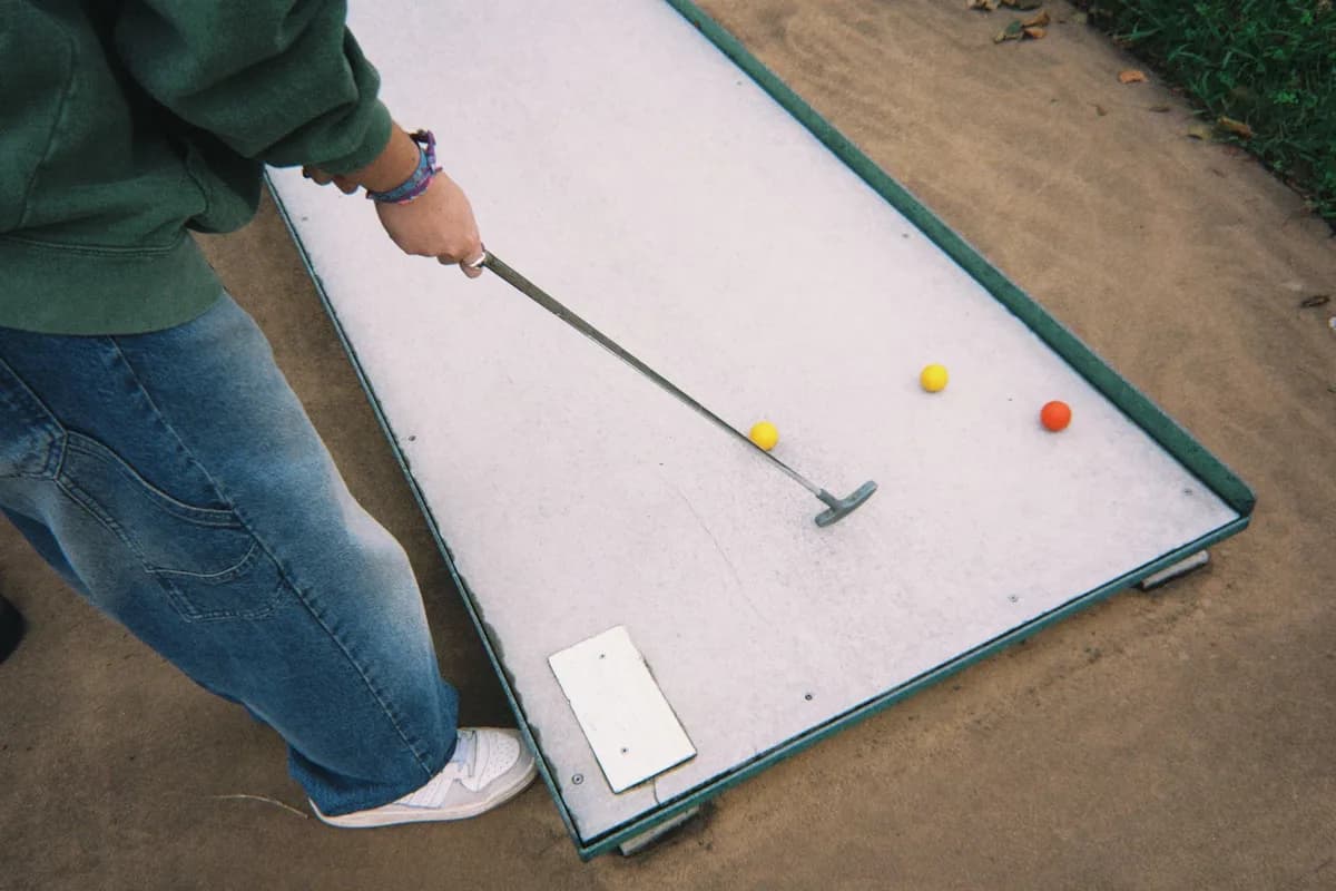 An adult playing mini golf outdoors with colored balls and putter on sand.