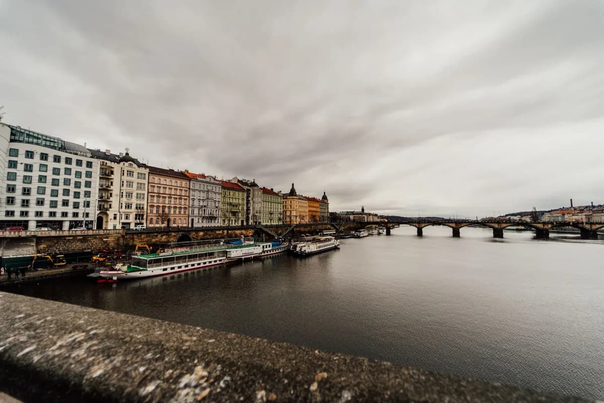 Scenic view of colorful buildings along the Vltava River in Prague with bridges under a cloudy sky.